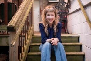 Press photo of Bronwyn Keith-Hynes a young woman with long wavy blonde hair and bangs wearing a navy blue tie-neck blouse and light wash jeans sitting on weathered wooden outdoor steps smiling warmly with hands clasped in front of her knees against a brick and concrete urban backdrop