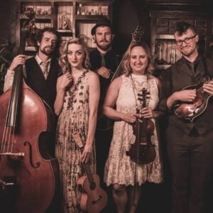 Pixie and The Partygrass Boys band press photo showing five members posed together in a warmly lit antique filled room with a man holding a double bass on the left a woman in a floral dress holding a ukulele a bearded man in a hat at center back a woman in a white lace dress holding a violin and a man holding a mandolin on the right