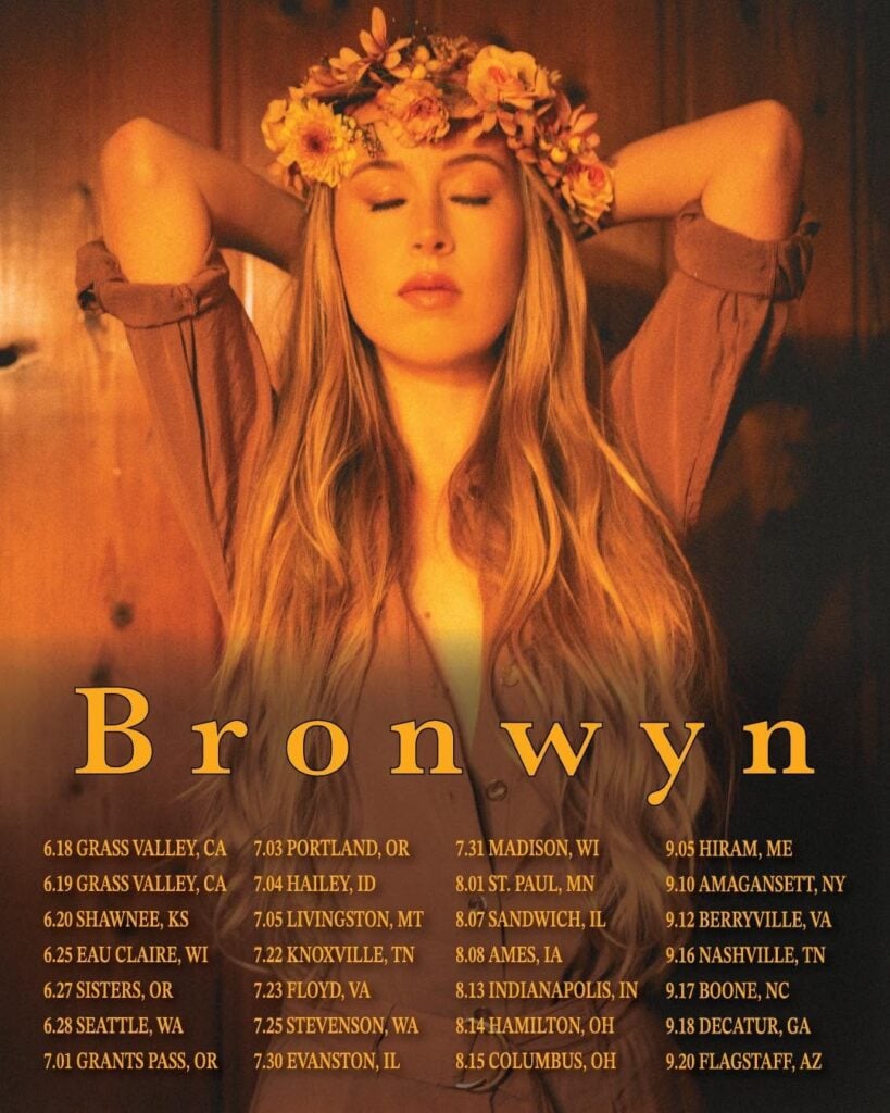 Tour poster for Bronwyn featuring a woman with long blonde hair wearing a floral crown and brown shirt with eyes closed and arms raised above her head bathed in warm golden light against a dark wood background with full tour dates listed below including Seattle June 28th 2026