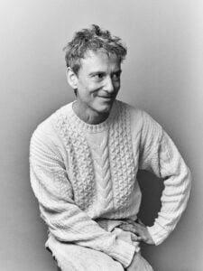 Black and white press photo of Teddy Thompson a middle aged man with short wavy salt and pepper hair wearing a chunky cable knit cream sweater sitting with one hand resting on his knee smiling softly and looking slightly off camera against a plain light grey background