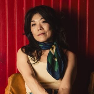 Press photo of BettySoo an Asian American woman with dark hair wearing a cream tank top and a colorful blue green and gold silk scarf around her neck sitting with arms crossed against a vivid red corrugated metal wall