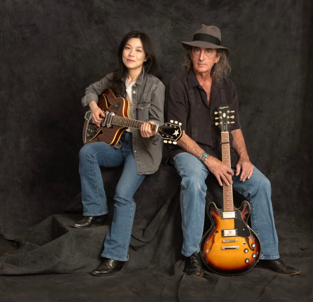 James McMurtry and BettySoo seated together for a press photo with BettySoo on the left holding a mandolin wearing a grey denim jacket and McMurtry on the right wearing a black shirt and grey fedora holding a sunburst electric guitar against a dark grey studio backdrop
