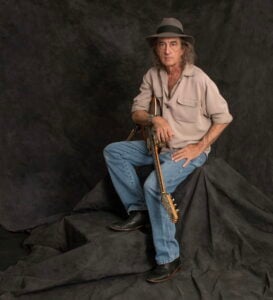 James McMurtry seated for a press photo wearing a tan button up shirt grey fedora and blue jeans with one hand resting on his knee and the other holding an acoustic guitar neck against a dark grey textured studio backdrop