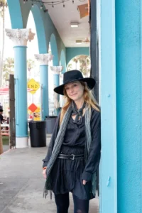 Eilen Jewell a woman with long wavy blonde hair wearing a wide brim black hat dark dress and fringed scarf standing and smiling beside a teal painted column under an arched outdoor walkway with palm trees visible in the background