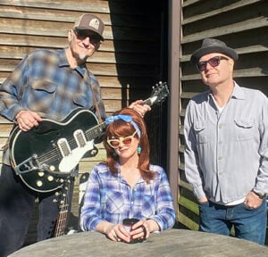 Southern Culture on the Skids band photo showing Rick Miller on the left holding a black electric guitar wearing a trucker hat and plaid shirt Mary Huff seated in the middle wearing a blue headscarf sunglasses and plaid shirt holding a coffee cup and Dave Hartman on the right wearing a fedora sunglasses and grey shirt standing outside against a wood plank wall