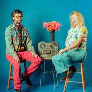 The Burning Hell duo Mathias Kom and Ariel Sharratt seated on wooden stools against a bright teal background with a reel to reel tape machine and vase of pink flowers between them Kom wearing a denim jacket covered in colorful patches and Sharratt wearing a floral print blouse and teal pants