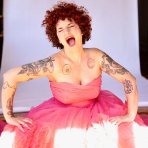 Press photo of Carsie Blanton a woman with curly red hair laughing with mouth wide open wearing a voluminous pink tulle dress with visible botanical and abstract tattoos covering both arms and shoulders sitting against a white draped backdrop with sunlight streaming across the bottom of the frame