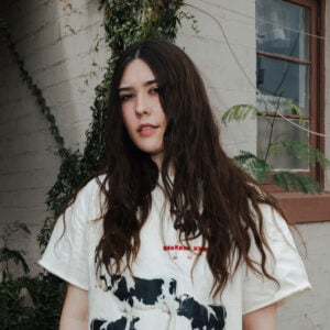Press photo of Sydney Sprague a young woman with long dark wavy hair wearing a white graphic t-shirt standing against a white brick wall with green ivy and plants in the background