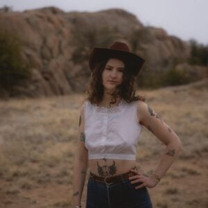 Press photo of Marley Hale a young woman with curly brown hair wearing a dark red wide brim cowboy hat white cropped button top and high waisted jeans with visible tattoos on both arms standing in a dramatic desert landscape with large boulders in the background