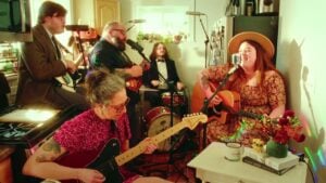 Emerson Woolf & The Wishbones performing an intimate live session in a warmly lit home kitchen with frontwoman Emerson Woolf singing into a vintage microphone while playing acoustic guitar wearing a floral dress and straw hat with four band members playing electric guitar mandolin and drums around her