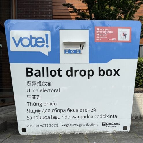 Blue and white ballot drop box with slot and multiple language printed on it.