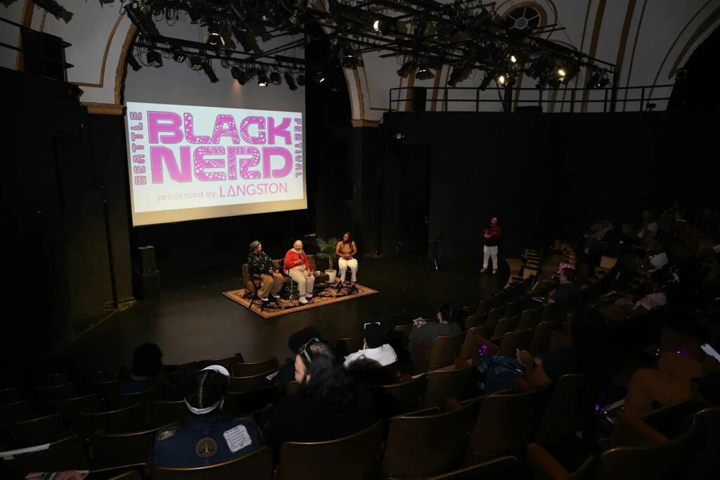 Wide angle interior shot of LANGSTON Theatre during Seattle Black Nerd Festival showing three panelists seated on a rug at the front of the darkened stage beneath a large projection screen displaying the Seattle Black Nerd Festival logo presented by LANGSTON in bold pink lettering with an audience of community members seated in wooden chairs filling the lower section of the theatre
