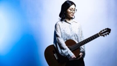 Young woman with glasses holds an acoustic guitar, looking thoughtfully aside in blue and lavender studio lighting; calm, artistic mood.