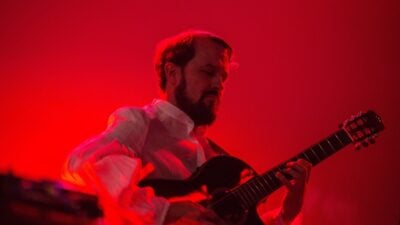Bearded musician plays electric guitar onstage under intense red lighting, his focused expression creating a dramatic, energetic concert mood.