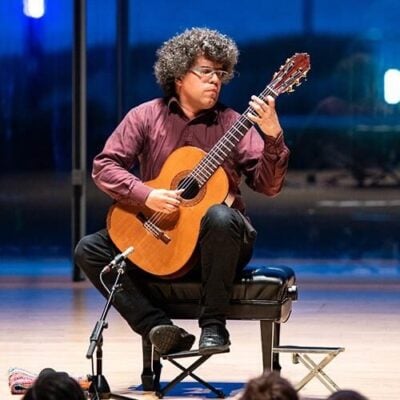 Curly-haired musician plays a classical guitar while seated onstage, focused and expressive under blue concert lights.
