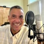 Smiling man in a home recording studio sits beside a microphone and pop filter, creating a warm, friendly podcasting scene.