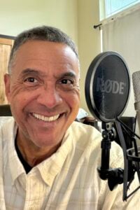 Smiling man in a home recording studio sits beside a microphone and pop filter, creating a warm, friendly podcasting scene.