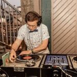 Focused DJ wearing headphones mixes vinyl records on dual turntables, with a central mixer, in a warm, vintage-style venue.