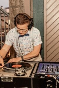 Focused DJ wearing headphones mixes vinyl records on dual turntables, with a central mixer, in a warm, vintage-style venue.