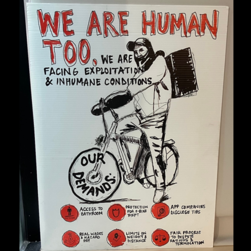 Photograph of a hand-drawn poster showing a delivery worker on a bicycle. Hand-drawn text reads "WE ARE HUMAN TOO, We are facing exploitation and inhumane conditions. Our demands: Access to bathroom, protection for e-bike theft, app companies disclose tips, real wages & hazard pay, limits on weight & distance, fair process to dispute ratings & termination."