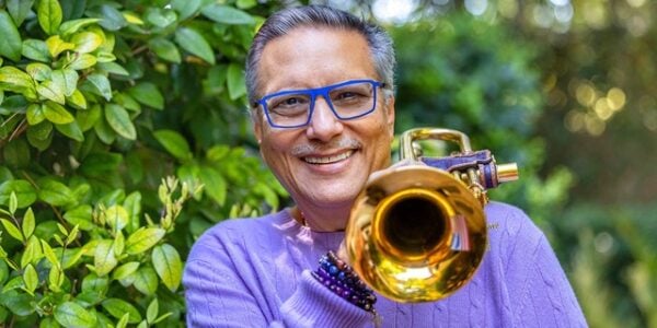 A smiling man in blue glasses and a purple sweater holds a shiny brass trumpet toward the camera, surrounded by lush green leaves in a cheerful outdoor scene.