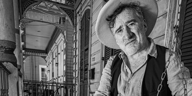 Black-and-white portrait of an older man in a wide-brimmed hat and vest, standing in an ornate corridor; his thoughtful gaze feels pensive.