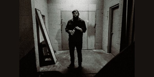 Grainy black-and-white image of a bearded man in dark clothing standing in a stark concrete hallway, hands near his chest, creating a tense, ominous mood.