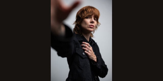 A red-haired person in a black outfit reaches toward the camera, their hand blurred in the foreground; the moody portrait feels bold and introspective.