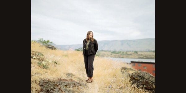 A woman in dark clothing stands on a rocky path amid golden grass, with distant hills and a cloudy sky creating a quiet, windswept mood.
