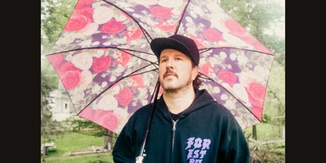 A mustached man in a black hoodie and wide-brimmed hat stands beneath a pink floral umbrella outdoors, looking thoughtful in the rain.