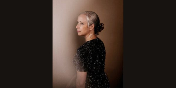 A woman with a silver bun and long earrings stands in profile, wearing a sparkling black dress against a softly lit brown backdrop; the mood is elegant and contemplative.