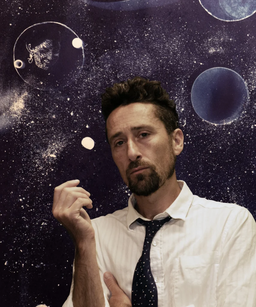 A pensive man in a white shirt and loosened dark tie raises one hand against a starry cosmic backdrop with glowing planets, creating a dreamy, contemplative mood.